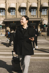 Smiling businesswoman with luggage holding smart phone while crossing street in city - MASF30611