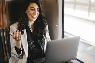 Lächelnde Geschäftsfrau führt einen Videogespräch am Laptop, während sie im Zug am Fenster sitzt - MASF30617