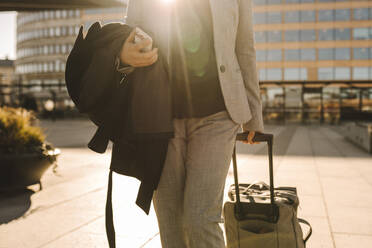 Midsection of businesswoman with smart phone pulling luggage while walking on sunny day - MASF30618