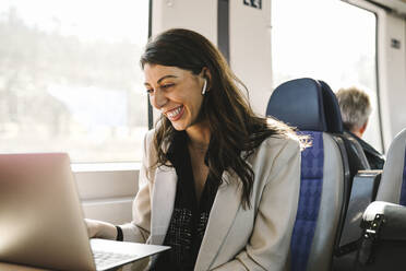 Happy businesswoman with in-ear headphones doing video call through laptop in train - MASF30627