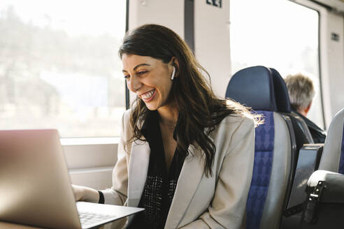 Glückliche Geschäftsfrau mit In-Ear-Kopfhörern bei einem Videogespräch über einen Laptop im Zug - MASF30627