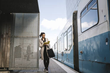 Businesswoman reading book while leaning at metal wall by train at railroad station - MASF30630