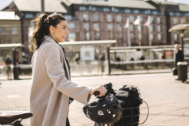 Seitenansicht einer Geschäftsfrau mit langem Mantel, die ein Fahrrad in der Stadt hält - MASF30635