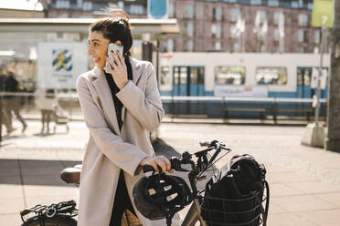 Businesswoman talking on smart phone standing with bicycle while looking back over shoulder in city - MASF30636