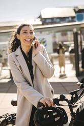 Glückliche Geschäftsfrau mit langem Mantel, die ein Fahrrad hält und mit einem Smartphone telefoniert - MASF30637