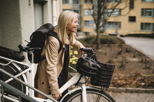 Seitenansicht einer lächelnden Geschäftsfrau mit Rucksack, die ein Elektrofahrrad hält - MASF30641