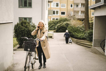 Happy businesswoman talking on smart phone while wheeling bicycle on footpath - MASF30645