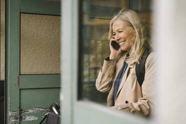 Smiling businesswoman talking on smart phone seen through glass window - MASF30648