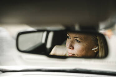 Businesswoman with in-ear headphones seen in rear-view mirror reflection of car - MASF30661