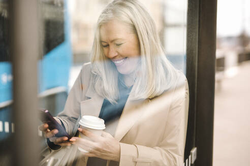 Glückliche Geschäftsfrau, die ein Smartphone benutzt und einen Einwegbecher hält, während sie sich an einer Bushaltestelle an ein Glas lehnt - MASF30664