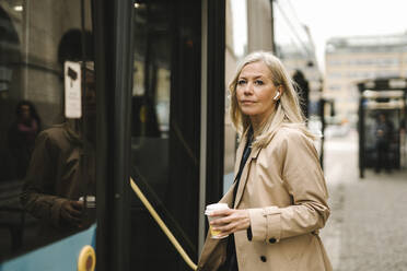 Businesswoman holding disposable cup while boarding bus in city - MASF30672