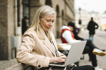 Blonde Geschäftsfrau, die einen Laptop benutzt und auf einer Bank in der Stadt sitzt - MASF30674