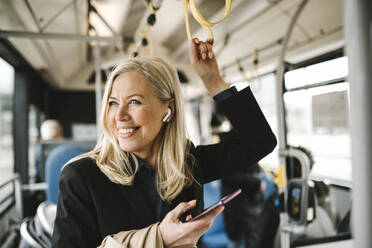 Happy businesswoman talking on smart phone holding grab handle while commuting through bus - MASF30677