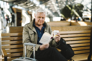 Ein älterer Geschäftsmann hält einen Einwegbecher und liest ein Buch, während er auf einer Bank in der Bahnhofsgaststätte sitzt - MASF30688