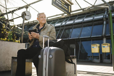 Geschäftsmann, der wegschaut und sein Smartphone in der Hand hält, während er auf einer Bank mit Gepäckstücken am Bahnhof sitzt - MASF30691