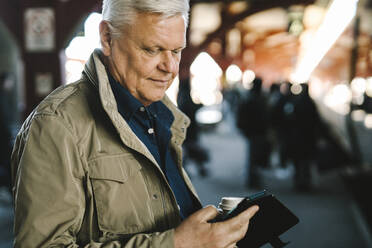 Geschäftsmann in Jacke und mit Smartphone am Bahnhof stehend - MASF30693