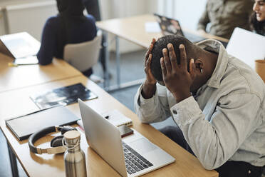 Müder männlicher Schüler mit dem Kopf in den Händen sitzt am Laptop auf dem Schreibtisch im Klassenzimmer - MASF30721