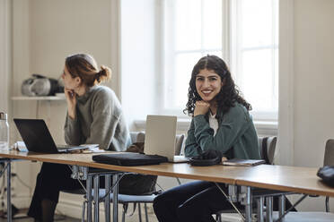 Porträt einer lächelnden Studentin mit Laptop, die mit einem Freund am Schreibtisch im Klassenzimmer sitzt - MASF30725