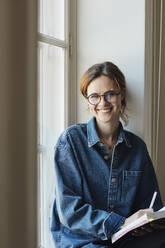 Portrait of happy female student with book by window - MASF30738