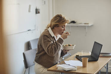 Professorin isst zu Mittag, während sie am Schreibtisch mit Laptop im Klassenzimmer sitzt - MASF30743