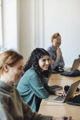 Glückliche Studentin mit Laptop, die inmitten von Freunden am Schreibtisch im Klassenzimmer sitzt - MASF30752