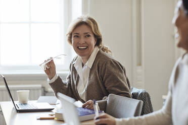 Happy female teacher holding eyeglasses looking at colleague in staffroom - MASF30754