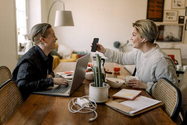 Glückliches lesbisches Paar, das sich bei der gemeinsamen Arbeit zu Hause ein Smartphone teilt - MASF30786