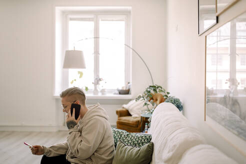 Seitenansicht einer Frau mit Schwangerschaftstestergebnissen, die über ihr Smartphone spricht, während sie auf dem Sofa im Wohnzimmer sitzt - MASF30800