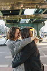Cheerful Lesbian couple embracing while standing on street under bridge in city - MASF30823