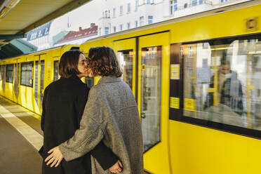 Lesbian couple kissing while standing by yellow train at railroad station - MASF30828