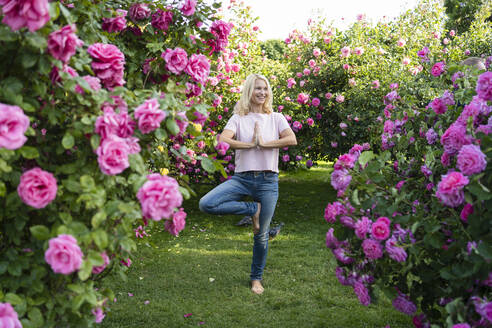 Happy woman doing tree pose yoga amidst rose garden - SVKF00228