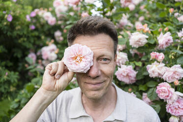 Lächelnder Mann hält im Garten eine Rose vor das Auge - SVKF00250