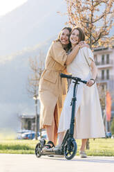 Glückliche Frauen, die sich gegenseitig umarmen und auf einem Motorroller im Park stehen - OMIF00815