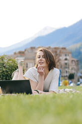 Smiling woman with laptop lying on grass at park - OMIF00821