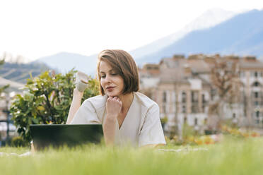 Frau mit Hand am Kinn und Laptop im Park liegend - OMIF00822