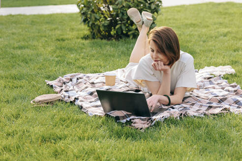 Woman with laptop lying on picnic blanket at park - OMIF00823