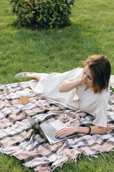 Frau mit Laptop auf Picknickdecke im Park liegend - OMIF00824