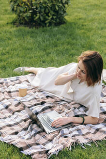 Woman using laptop lying on picnic blanket at park - OMIF00824