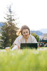 Woman with tousled hair using laptop lying at park - OMIF00827
