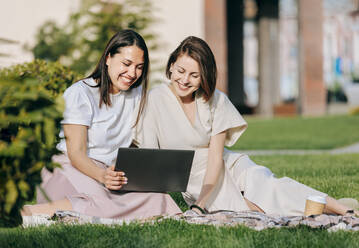 Glückliche Freundinnen teilen sich einen Laptop und sitzen im Park an einem sonnigen Tag - OMIF00828