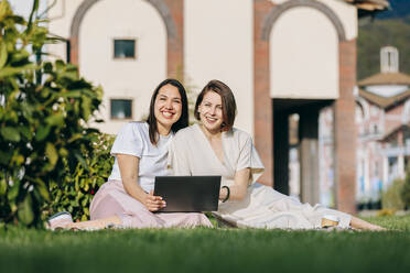 Glückliche Freundinnen sitzen mit Laptop im Park an einem sonnigen Tag - OMIF00829