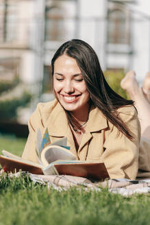 Happy beautiful woman reading book lying at park on sunny day - OMIF00831