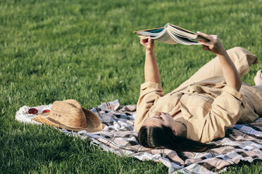 Junge Frau liest auf einer Picknickdecke im Park liegend ein Buch - OMIF00834