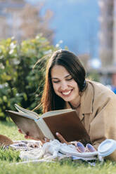 Glückliche junge Frau mit dunklem Haar, die im Park liegend ein Buch liest - OMIF00836