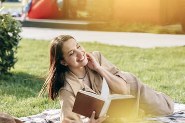 Lächelnde Frau mit geschlossenen Augen, die ein Buch hält und im Park genießt - OMIF00838