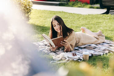 Lächelnde junge Frau, die auf einer Picknickdecke im Park liegt und ein Buch liest - OMIF00840