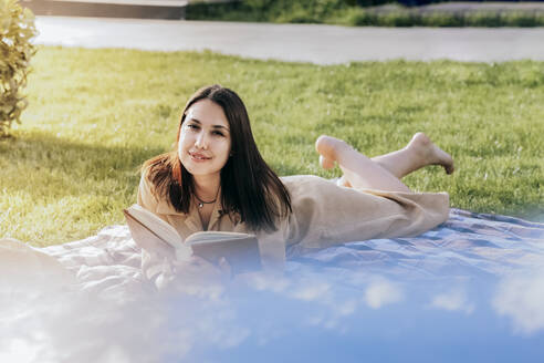 Beautiful woman lying with book on picnic blanket at park - OMIF00841