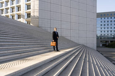 Mature businessman holding bag standing on staircase in front of building - OIPF01972