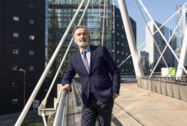 Mature businessman with hand in pocket standing by railing on sunny day - OIPF01998