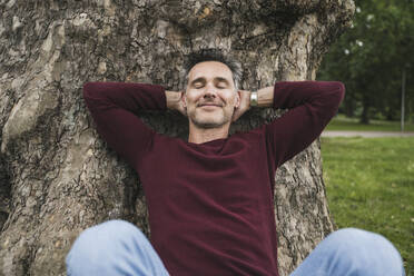 Smiling mature man with hands behind head leaning on tree trunk - UUF26241